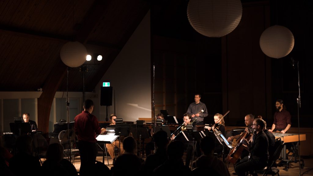 A photo of musicians performing in a semi-lit concert space. Large white paper spheres hang from the ceiling.